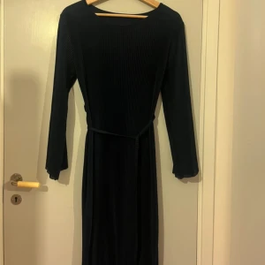 Mörkblå plisserad långklänning - Säljer en elegant mörkblå långklänning med plisserat tyg och lång ärm. Klänningen har ett smalt bälte i midjan och en mjuk, vågig kant nertill. Perfekt för dig som gillar stilrena och klassiska plagg.