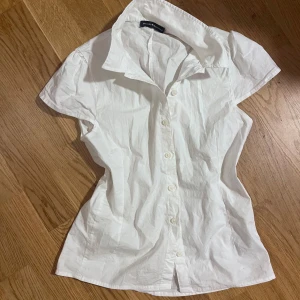Vit kortärmad figursydd skjorta från Brandy Melville - Säljer en klassisk populär vit skjorta som är figursydd från Brandy Melville med korta ärmar och knappar framtill. Skjortan har en feminin passform och är tillverkad i ett lätt bomullsmaterial. Perfekt till sommaren och enkel att styla till olika outfits. Aldrig använt. Nypris runt 500kr
