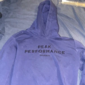 Blå hoodie från Peak Performance - Säljer en blå hoodie från Peak Performance med broderad logga på bröstet. Tröjan har huva och är tillverkad i mjukt material, perfekt för en avslappnad stil.