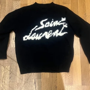 Svart stickad tröja från Saint Laurent - Svart stickad tröja med vit Saint Laurent-logga i grafisk stil över bröstet. Klassisk rund halsringning och långa ärmar. Perfekt för dig som gillar statement-plagg och exklusiva märken.