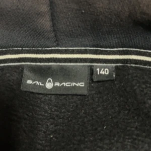 Svart hoodie från Sail Racing med dragkedja - Mörkgrå/svart hoodie från Sail Racing med hel dragkedja framtill och huva. Tröjan har en bröstficka med dragkedja och logga på bröstet. Perfekt för kyliga dagar och enkel att matcha med det mesta. Den är lite sol blek men ändå bra