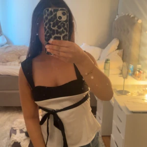 Vit och svart tubtopp med rosett - Säljer en snygg vit tubtopp med svarta detaljer och breda axelband. Toppen har en svart rosett i midjan och svarta kanter upptill. Perfekt till sommaren och passar till både jeans och kjol.