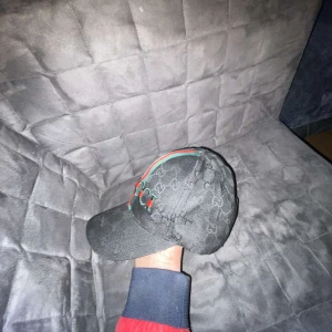 Svart keps från Gucci med logga - Svart keps från Gucci med diskret GG-mönster över hela kepsen och broderad logga framtill. Kepsen har gröna och röda ränder längs ovansidan och justerbart spänne bak. Snygg och klassisk design.