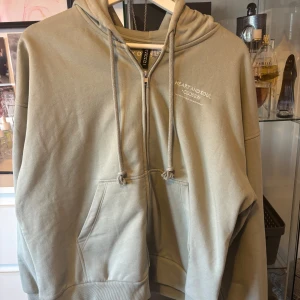 Grön hoodie med dragkedja från H&M Divided - Snygg ljusgrön hoodie från H&M Divided med dragkedja och huva. Tryck med texten 'JOY DIVISION' på ena ärmen och liten vit text på bröstet. Mjuk bomullskänsla och känguruficka framtill. Ljuset är lite dåligt på bilderna, så tröjan ser mer gul ut på bild än i verklighet.