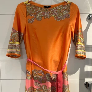 Orange paisleymönstrad klänning från H&M - Färgstark orange klänning från H&M med paisleymönster i rosa, gult och vitt. Klänningen har trekvartsärm, bälte i midjan och är tillverkad i ett mjukt, glansigt material. Perfekt för dig som gillar unika mönster och färg.