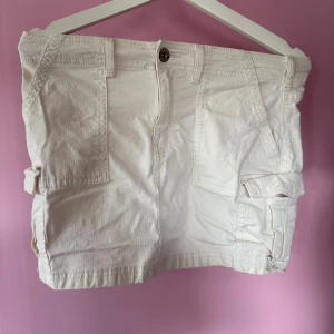 Vit cargokjol från Bershka - Vit cargokjol från Bershka med flera fickor och knappstängning framtill. Kjolen har en kort längd och är tillverkad i bomull, perfekt för en avslappnad och trendig stil.