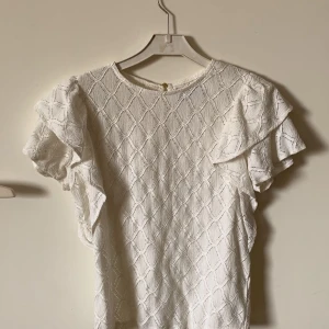 Vit blus med volang från Holly & Whyte - Säljer en vit blus från Holly & Whyte med strukturmönster och volangdetaljer på ärmarna. Blusen har korta ärmar och en liten knapp i nacken. Perfekt för en fräsch och stilren look.