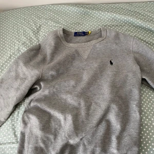 Grå sweatshirt från Polo Ralph Lauren - Klassisk grå sweatshirt från Polo Ralph Lauren med rund halsringning och broderad logga på bröstet. Tröjan har långa ärmar och är tillverkad i mjukt bomullsmaterial. Perfekt för en avslappnad och stilren look. Helt nytt skick riktigt skön