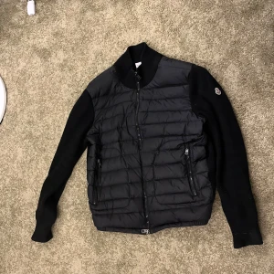 Svart hybridjacka från Moncler - Säljer en svart Moncler-jacka med quiltad front i syntet och stickade ärmar och rygg. Jackan har dragkedja framtill, två fickor med dragkedja och Moncler-logga på ärmen. Perfekt för lager-på-lager under kyliga dagar.