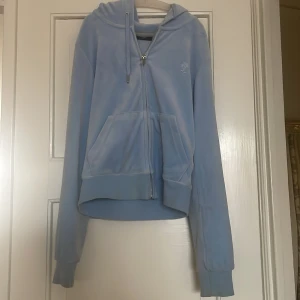 Ljusblå velour hoodie från Juicy Couture - Säljer en ljusblå hoodie i mjuk velour från Juicy Couture. Jackan har dragkedja, huva med snörning och fickor framtill. Ikonisk JC-logga broderad på bröstet. Perfekt för en avslappnad och trendig look.