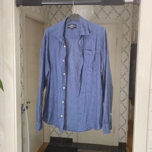 Blå linneskjorta med vita knappar - Snygg blå skjorta i linne från Hampton Republic  med klassisk krage och vita knappar. Skjortan har en bröstficka och lång ärm, perfekt för en avslappnad men stilren look.