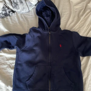 Mörkblå hoodie från Polo Ralph Lauren - Mörkblå hoodie från Polo Ralph Lauren med dragkedja och huva. Tröjan har två fickor framtill och en röd broderad logga på bröstet. Perfekt för en avslappnad stil. 150cm storlek 