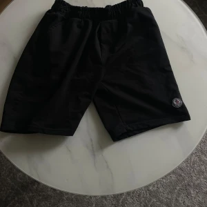 Svarta shorts från Moncler - Svarta shorts från Moncler med elastisk midja och diskret Moncler-logga på vänster ben. Klassisk passform med två fickor bak och mjukt material som känns bekvämt. Perfekta för sommaren.