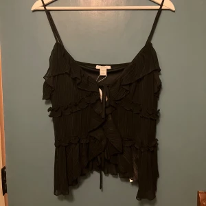 Svart trendig topp - Säljer en svart trendig topp från NewYorker med smala axelband, flera volangdetaljer framtill och knytning fram. 