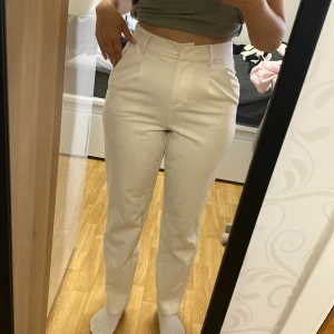 Vita kostymbyxor med hög midja - Säljer ett par stilrena vita kostymbyxor med hög midja och raka ben. Byxorna har bälteshällor och stängs med dragkedja och knapp. Perfekta för en clean och fräsch look.