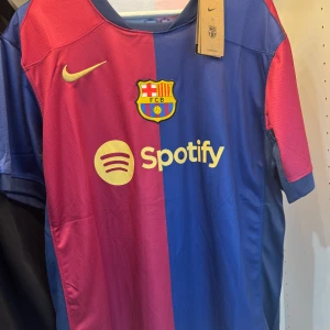 FC Barcelona matchtröja 2023/24 - Säljer en officiell FC Barcelona fotbollströja från Nike med Spotify-logga på bröstet. Tröjan är halva röd och halva blå, med klubbmärke och Nike-logga framtill. Kortärmad modell i lätt och ventilerande material, perfekt för match eller träning.