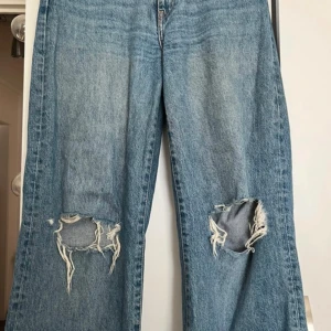 Blå slitna jeans med hål på knäna - Säljer ett par blå jeans med bred passform och stora slitna hål på båda knäna. Jeansen har klassisk femficksdesign och är tillverkade i kraftigt denimtyg. Perfekta för en avslappnad och trendig look. Säljer samma på vinted så kontakta gärna innan ni trycker på ”köp”!