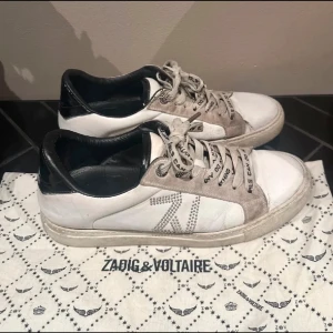 Vita sneakers från Zadig & Voltaire - Snygga vita sneakers med grå mockadetaljer och snörning. Skorna har en glittrig stjärndetalj på sidan och text på skosnörena. Perfekta för en trendig look. Tyvärr är de lite slitna som synd på bild tre men inget man tänker på 