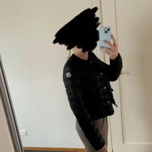 Svart dunjacka från Moncler - Snygg svart dunjacka från Moncler med knappar framtill. Jackan har en normal passform och är perfekt för kyligare dagar. Den är långärmad och har Monclers logga på ärmen.