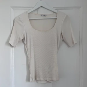 Beige ribbad topp från Wera - Säljer en stilren vit ribbad topp från Wera. Toppen har en rundad halsringning och korta ärmar, perfekt för en minimalistisk look. Passar bra till både jeans och kjol.