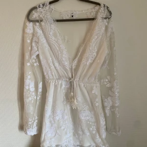 Vit spetsklänning med paljetter - Säljer en vacker vit spetsklänning med paljetter och v-ringning. Klänningen har långa ärmar och ett knytband i midjan för en fin passform. Perfekt för speciella tillfällen!