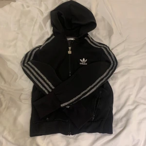 Svart huvtröja / kofta från Adidas - Säljer en svart huvtröja från Adidas med dragkedja och vita detaljer. Tröjan har långa ärmar med ett mönster längs sidorna och en broderad logga på bröstet. Den har även fickor med dragkedja framme på båda sidor. 