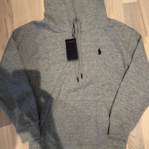 Grå hoodie från Polo Ralph Lauren S - Säljer en stilren grå hoodie från Polo Ralph Lauren med det klassiska logotypen broderad på bröstet. Tröjan har en justerbar huva med snören och en praktisk magficka. Perfekt för en avslappnad look.