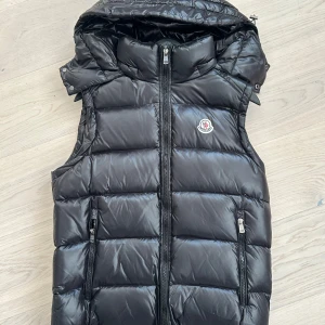 Svart dunväst från Moncler - Snygg svart dunväst från Moncler med dragkedja och avtagbar huva. Västen har en quiltad design och två praktiska fickor med dragkedja framtill. Perfekt för kyligare dagar när du vill hålla stilen. Storlek S/M. Avtagbar luva