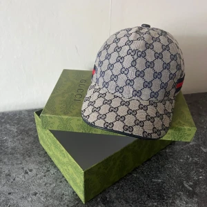 Mönstrad keps från Gucci - Snygg keps från Gucci med det ikoniska GG-mönstret i beige och blått. Kepsen har en röd och blå randdetalj på sidan och är tillverkad i Italien. Perfekt för att ge din outfit en lyxig touch.