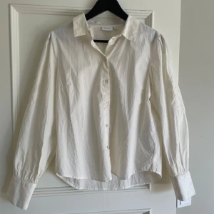 Vit blus med broderade detaljer - Elegant vit blus från Vila med långa ärmar och knappar framtill. Blusen/sjortan har vackra broderade detaljer på ärmarna som ger en stilren look. Den är crem färgade/vit