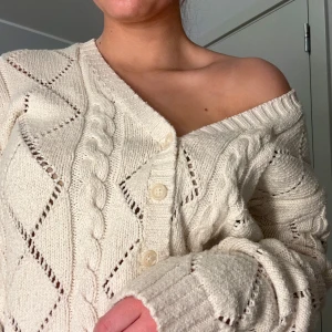 Beige stickad kofta från Brandy Melville - Säljer en mysig beige stickad kofta från Brandy Melville. Koftan har ett fint hålmönster och knappar framtill. Den är offshoulder och har ribbade kanter vid ärmslut och nederkant. Perfekt för en avslappnad stil.