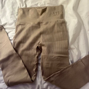 Beige ribbade leggings från Stronger - Snygga beige leggings från Stronger med ribbad textur och hög midja. Perfekta för träning eller en avslappnad dag. De har en stilren design med diskreta detaljer och är superbekväma att bära. Helt nya och aldrig andvända.