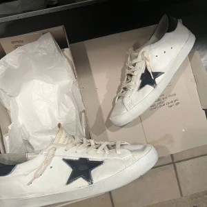 Golden goose skor - Snygga vita sneakers med en blå stjärna på sidan och blå detaljer vid hälen. Skorna har snörning och en rund tå. Perfekta för en avslappnad stil. Har lagt på lite ny fräsch färg på dom. Priser kan diskuteras vid snabb affär. Skriv Järna om du har funderingar!