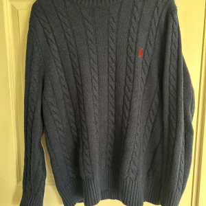 Ralph Lauren tröja - Säljer en snygg blå stickad tröja från Polo Ralph Lauren med kabelstickat mönster och röd broderad logga på bröstet. Tröjan har långa ärmar och ribbade muddar vid ärmslut och nederkant. Perfekt för kyliga dagar!
