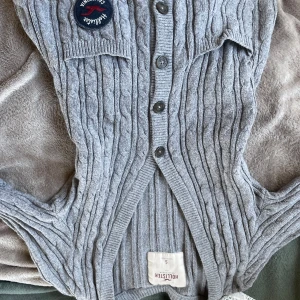 Grå ribbad väst från Hollister - Snygg grå ribbad väst från Hollister med knappar framtill och ett märke på bröstet. Perfekt för lager-på-lager-stil och att hålla sig varm under kyliga dagar.