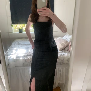 Svart långklänning från Bubbleroom - Elegant svart långklänning från Bubbleroom med smala axelband och öppen rygg med snörning. Klänningen har en slits framtill och är perfekt för festliga tillfällen. Materialet ger en lyxig känsla, perfekt till bal eller andra festliga tillfällen. Endast använd 1gång.