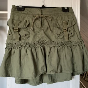 Olivgrön kjol med fickor - Snygg olivgrön kjol med stora fickor och knytband i midjan. Kjolen har volangdetaljer och en avslappnad passform. Perfekt för en avslappnad stil.