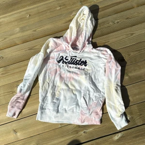 Flerfärgad hoodie från Hollister - Säljer en snygg flerfärgad hoodie från Hollister med tryck på bröstet. Hoodien har en klassisk känguruficka och justerbar huva med dragsko. En M men liten i storlek så lägger in den som en S. Perfekt för en avslappnad stil.