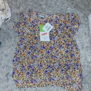 Blommig topp - Säljer en färgglad blommig topp som har ett livligt mönster i lila, orange och vit med elastiska detaljer vid ärmslut och nederkant. Perfekt för sommarens varma dagar! Det står tyvärr inget pris då det bara är halva prislappen kvar.