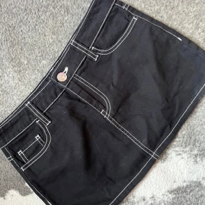 Svart kortkjol med kontrastsömmar - Snygg svart mini kjol med vita kontrastsömmar och praktiska fickor fram och bak. Kjolen har en knappdetalj i silver och en klassisk jeansstil. Perfekt för en trendig look.