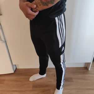 Svarta träningsbyxor från Adidas - Snygga svarta träningsbyxor från Adidas med de klassiska vita ränderna längs sidorna. Byxorna har en tight passform och är perfekta för träning. De har en elastisk midja för extra komfort.