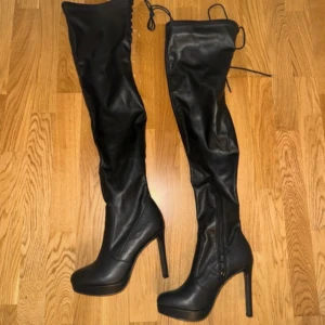 Svarta overknee stövlar med klack - Snygga svarta overknee stövlar i syntetmaterial med hög klack och dragkedja på sidan. Perfekta för en stilren look.