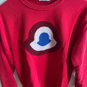 Röd tröja från Moncler - Säljer en snygg röd tröja från Moncler med ett stort broderat märke på framsidan i blått och vitt. Tröjan har en klassisk rund hals och är perfekt för en stilren look. Passar bra till både jeans och chinos.