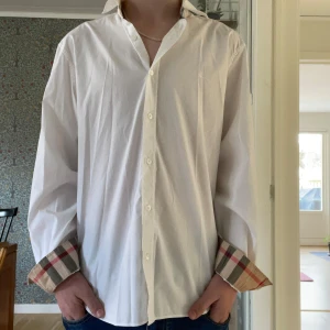 Vit skjorta från Burberry - Säljer en stilren vit skjorta från Burberry med klassiska rutiga detaljer på ärmslut och krage. Skjortan har långa ärmar och knappar framtill. Perfekt för en elegant look. Passar som S-M.