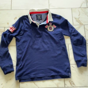 Blå pikétröja från Robbie Moor - Säljer en snygg blå pikétröja från Robbie Moor med broderad logga och brittisk flagga på ärmen. Storlek L men den passar som M. Plagget har inga defekter. Kom gärna privat för frågor m.m.🙌