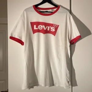 Vit t-shirt från Levi's med röd logga - Säljer en klassisk vit t-shirt från Levi's med röd logga på bröstet. T-shirten har röda kanter vid ärmar och hals, vilket ger en snygg kontrast. Perfekt för en avslappnad stil. Tillverkad i Turkiet.
