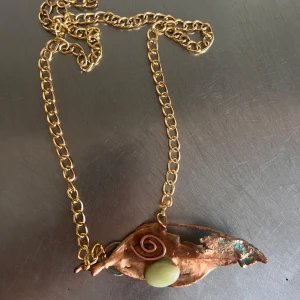 Unikt halsband med bladmotiv - Ett unikt halsband med ett bladformat hänge i koppar och grönt, dekorerat med en spiral och en grön sten. Kedjan är i guldton och ger en elegant touch. Perfekt för den som gillar naturinspirerade smycken.
