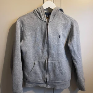 Grå hoodie från Ralph Lauren - Säljer en klassisk grå hoodie från Ralph Lauren med dragkedja och fickor framtill. Den har en broderad logga på bröstet och är tillverkad i mjukt material för extra komfort. Perfekt för en avslappnad stil.