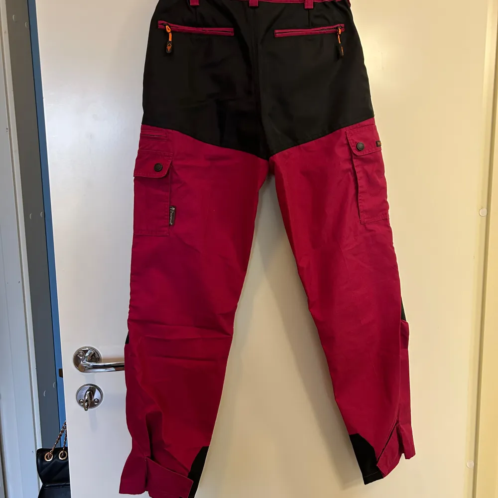 Snygga och funktionella friluftsbyxor från Pinewood i rosa och svart. Byxorna har flera praktiska fickor med knappar och dragkedjor, perfekt för utomhusaktiviteter. De är designade för att vara slitstarka och bekväma, med förstärkningar på knän och bakdel.. Farkut & Housut.