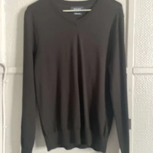 Svart merinoulltröja från Riley - Säljer en stilren svart tröja från Riley i mjuk merinoull. Tröjan har en v-ringad design och långa ärmar, perfekt för kyligare dagar. Passar både till vardags och mer uppklädda tillfällen. Ett måste i garderoben för den som gillar klassisk stil.